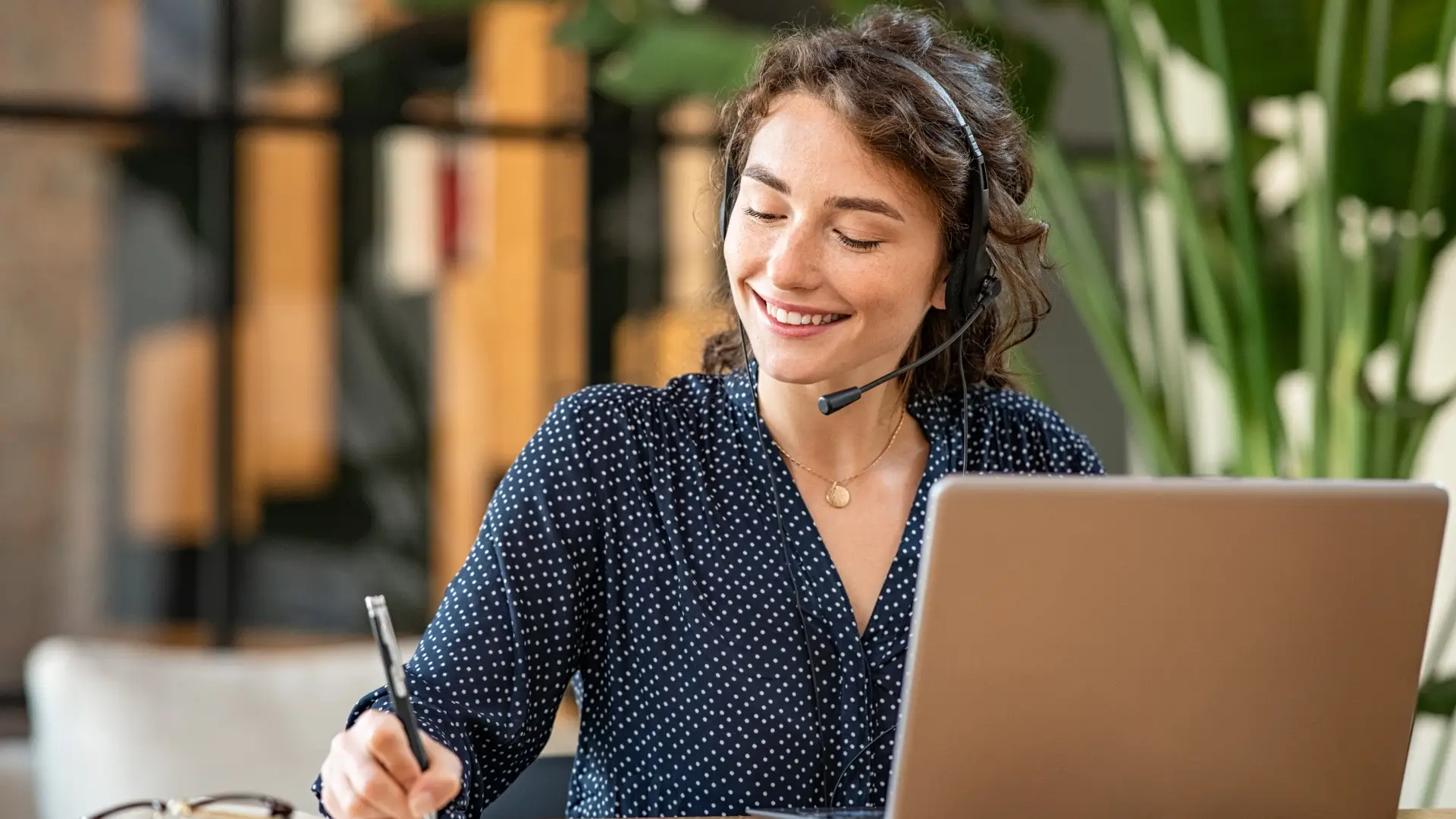 Symptisch lächelnde Frau mit Headset vor einem Laptop, schreibt sich etwas mit Kugelschreiber auf