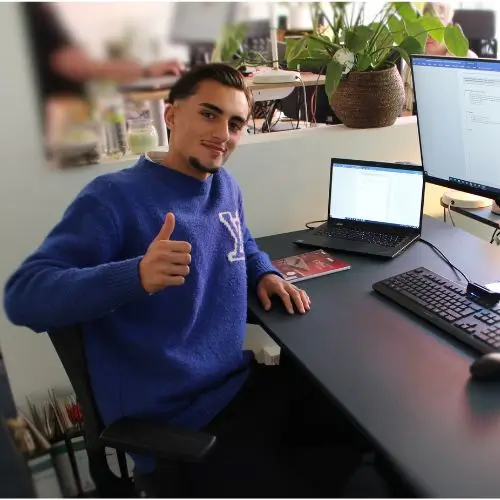 Auszubildender Diyar am Schreibtisch, guckt freundlich und zeigt einen Dauemn nach oben.